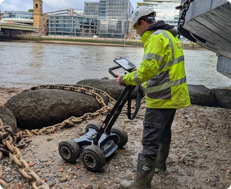 A surveyor using a GPR device near a riverbank to check for structural issues behind a masonry retaining wall