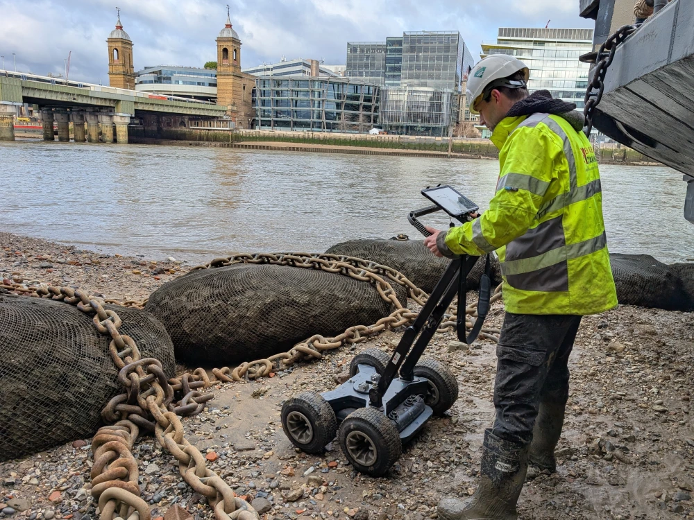 A surveyor using a ground penetrating radar device near a riverbank to check for structural issues behind a masonry retaining wall.